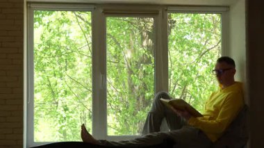 Sarı kapüşonlu ve gözlüklü genç bir adam pencere pervazında oturuyor, kitap okuyor ve sayfaları çeviriyor. Pencerenin dışında yeşil ağaçlar görülebilir, sakin ve rahat bir atmosfer yaratırlar. 4k
