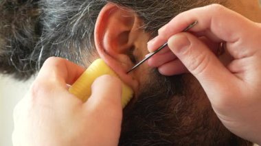 Bir adamın dikkatle bir başkasının kulağını steril bir iğneyle delmesi. Temiz aletler kullanılarak düzenlenmiş bir prosedür. Güvenli bir ev ortamında kulağına piercing takmanın göstergesi.