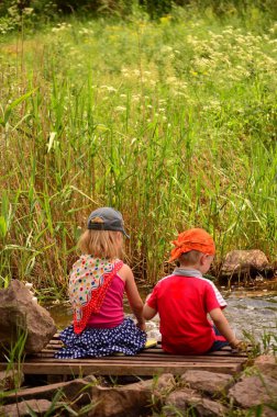 Nehir kenarındaki küçük ahşap bir köprüde oturan iki çocuğun dikey fotoğrafı. Etrafı uzun otlar ve kayalarla çevrili. Çocukluk ve doğa bağlantısını gösteren huzurlu yaz manzarası.