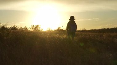 4K videosunda sırtında sırt çantası olan bir çocuk bozkırda gün batımına doğru yürüyor. Çocukluk macerası, seyahat, özgürlük ve yeni başlangıçların sembolü