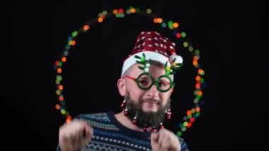 Sakalında oyuncaklar, arka planında yeni yıl ışıkları ve boynuz şeklinde yeni yıl gözlüğü olan Noel Baba şapkalı genç bir adam. Kutlamada dans edip eğleniyoruz.