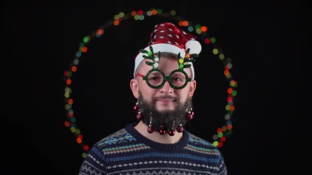 サンタクロースの帽子の中の若い男は 新年の光を背景に 角の形をした新年の眼鏡で彼のひげにおもちゃをつけています メアリー クリスマスを叫びクリスマスツリーを手に — ストック動画