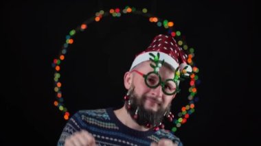 Sakalında oyuncaklar, arka planında yeni yıl ışıkları ve boynuz şeklinde yeni yıl gözlüğü olan Noel Baba şapkalı genç bir adam. Kutlamada dans edip eğleniyoruz.