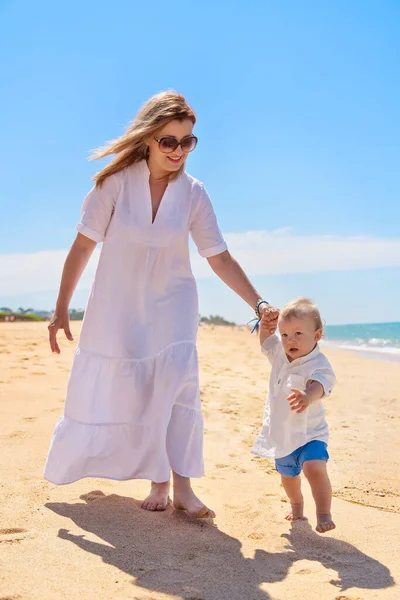 Caucasian mom walks with a happy one-year-old son along the sea beach in the summer, taking first steps. Concept of happy childhood and motherhood. High quality photo