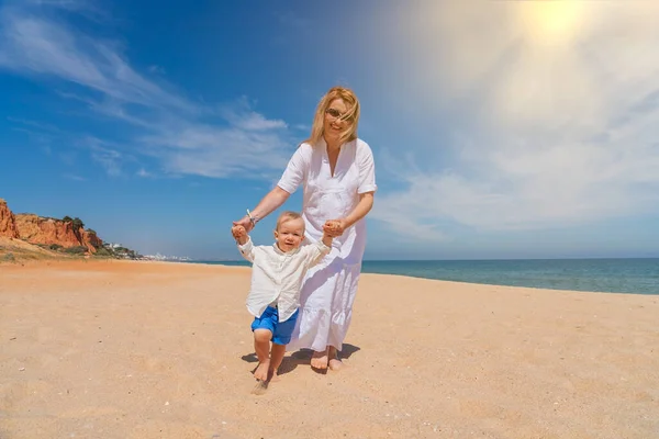 Young caucasian mother with a happy son walks along the beach in summer with bright sun and turquoise sea. Concept of happy childhood and motherhood