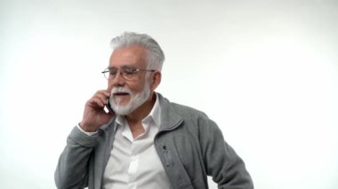 Portrait of an elderly retired man in glasses talking on a mobile phone on a white background. Gray-haired grandfather with a beard posing on an isolated studio background. Slow motion. High quality