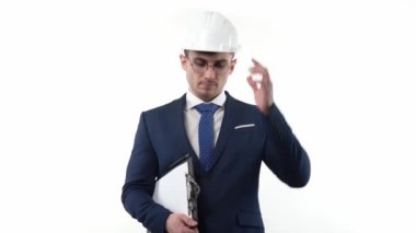 A man businessman in a suit with glasses in a protective helmet on a white background, thinking about solving a problem, holds a notepad in his hands for writing