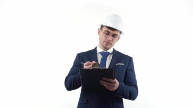 Male businessman in a suit with glasses in a protective helmet thinks and makes planning notes with a pen in a notebook. On a white background. High quality FullHD footage