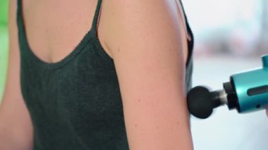 A young female athlete uses a modern massage gun for therapeutic relaxation of the triceps arm. The concept of sports recovery after training and rejuvenation