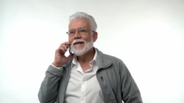 Portrait of an elderly retired man in glasses talking on a mobile phone on a white background. Gray-haired grandfather with a beard posing on an isolated studio background. Slow motion. High quality