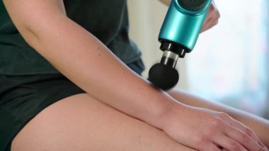A young female athlete uses a modern massage gun for therapeutic relaxation of the arm and forearm. The concept of sports recovery after training and rejuvenation. High quality 4k footage