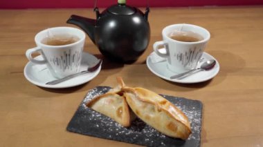 Appetizing fresh empanadas on a stone tray close-up and cups with a teapot in the background. High quality FullHD footage