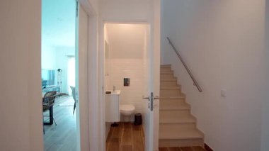 Beyaz metro fayanslarının olduğu kompakt modern bir banyo, duvara monte edilmiş bir tuvalet, dolaplı küçük beyaz bir lavabo, bir ayna, bir çöp kutusu ve merdiven altındaki ahşap döşeme..