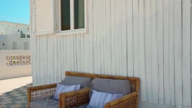 Gri minderli ve çizgili yastıklı iki şık hasır sandalye güneşli teras dekoratif fenerinde beyaz ahşap duvara karşı duruyor.