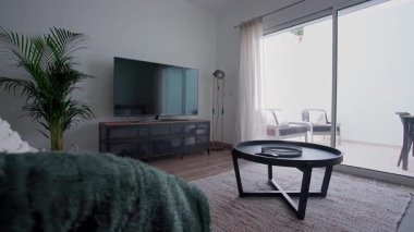 Siyah yuvarlak sehpası olan minimalist bir oturma odası, büyük bir televizyon ve açık hava avlusuna açılan kapalı bir bitki. Yumuşak doğal ışık boşluğu doldurur modern ev konforu ve iç tasarım kavramı