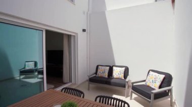 Siyah yastıklı minimalist bahçe verandası çiçek desenli yastıklar ve beyaz duvarlara karşı ahşap yemek masası sıcak ve davetkar bir atmosfer yaratıyor.