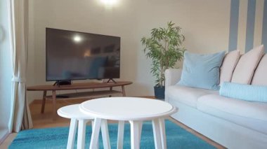 Bej koltuklu, yumuşak mavi yastıklı ve çizgili aksanlı duvarlı modern oturma odası. Büyük bir televizyon ahşap bir stantta otururken, bir saksı bitkisi rahat ve zarif bir ev iç mimarisi kavramına sahiptir.