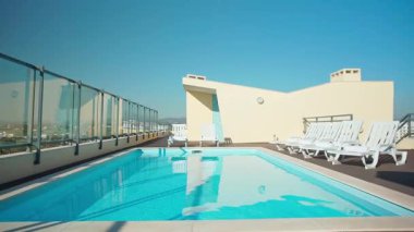 Beyaz şezlonglar ve cam bariyerlerle çevrili kristal berraklığında mavi suyu olan çatı yüzme havuzu parlak güneşli bir gökyüzü altında panoramik bir manzara sunuyor. Lüks rahatlama atmosferi