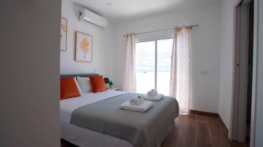 Minimalist yatak odası, düzgün yapılmış portakal renkli yastıklar ve çerçevelenmiş botanik baskılar desenli perdeleri olan büyük bir pencere..