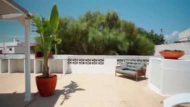 Geniş çatı terasında gri yastıklı hasır bir kanepe, dahili bir barbekü alanı, dekoratif beyaz duvarlar, yemyeşil ve arka planda açık mavi bir gökyüzü var..