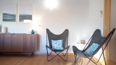 Minimalist oturma odası, iki modern kelebek koltuğu, ahşap bir büfe ve mavi dekoratif aksanlı. Hafif ahşap döşeme ve tarafsız duvarlar rahat ve çağdaş bir atmosfer yaratıyor.