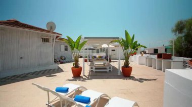 Geniş bir teras, beyaz güneş şezlongları, pergola altında ahşap bir masa ve büyük yeşil saksı bitkileri. Açık hava Akdeniz atmosferini güçlendirir. Açık hava rahatlama kavramı