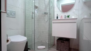 Modern bir banyoda cam kaplı fayanslı bir duş, yuvarlak aynalı yüzen bir kibir, beyaz bir tuvalet, dekoratif tuvalet malzemeleri ve dokuma sepetleri bulunur..