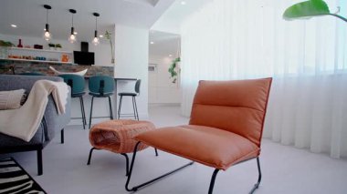 Çağdaş bir yaşam alanı. Kahverengi deri bir şezlong, dokunmuş bir şilte ve modern bir mutfak manzarası, kolye ışıklandırması ve zarif bir dekor olan parlak gri bir kanepe..