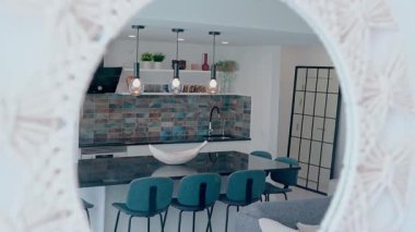 Çağdaş bir mutfak, parlak siyah bir tezgah adası, mavi döşemeli bar taburesi, şık bir döşeme kaplaması, ve dekoratif bir aynaya yansıyan kolye ışığı..