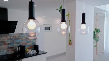 Pürüzsüz cam küreler ve siyah eşyalar ile üç modern kolye mutfak tezgahının üzerinde asılı, beyaz dolaba karşı sıcak ışık yansıtıyor ve seramik kaplama.