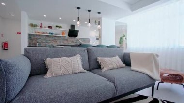 Şık oturma odasında dekoratif yastıklı gri kumaş bir kanepe, beyaz bir atış, siyah ve beyaz çizgili bir halı, saksı bitkileriyle yuvarlak siyah bir kahve masası ve sıcak bir ışıklandırma bulunuyor..