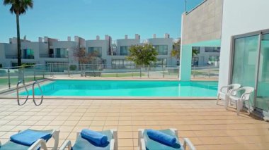 Mavi havlular, cam çitler ve parlak mavi gökyüzünün altındaki modern beyaz villalar ile çevrili, kristal berrak suyla dolu lüks bir açık yüzme havuzu..