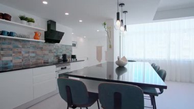 Beyaz dolaplı çağdaş bir açık konsept mutfak, siyah bir tezgah, mavi tonlu döşeme kaplaması, ve kolye ışıklı ve bar tabureli geniş bir yemek alanı..