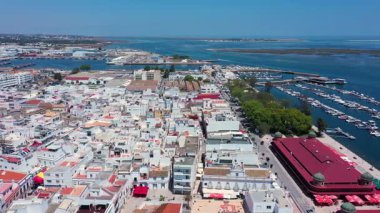 Portekiz 'in Olhao şehrinin yüksek açılı insansız hava aracı görüntüsü. Geleneksel beyaz binalar, kırmızı çatılı Olhao Pazarı, hareketli bir marina ve açık mavi gökyüzü altındaki Ria Formosa' nın çarpıcı suları..
