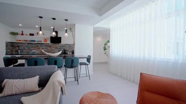 Kahverengi deri bir şezlong, dokunmuş bir şilte ve kolye aydınlatması ve açık bir mutfak ile tamamlanmış rahat fırlatmalı gri bir kanepe içeren çağdaş yaşam alanı..