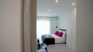 Düzgün yapılmış bir yatak odası, canlı pembe yastıklar, koyu renk döşemeli yatak başlığı, yumuşak ışıklandırma, gömme gardıroplar, siyah bir sandalye ve doğal ışığı filtreleyen katıksız perdeler..