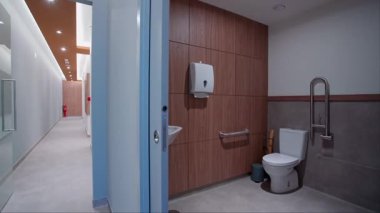Beyaz tuvaleti, paslanmaz çelik parmaklıkları, duvara monte edilmiş bir kağıt dağıtıcısı ve parlak ışıklandırmalı parlak bir koridorun bitişiğindeki ahşap duvar panelleri olan modern erişilebilir bir tuvalet..