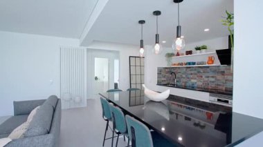 Parlak siyah bir adası olan modern bir açık konsept mutfak, kolye ışıkları, mavi kartonpiyer kaplama, beyaz dolaplar, bar tabureleri ve dekoratif elementlerle dolu raflar..