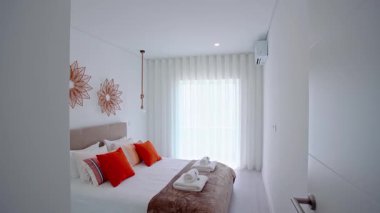 Bej döşemeli, turuncu yastıklı ve dekoratif duvar resimli parlak bir yatak odası. Perdeler geniş bir pencereyi kaplıyor ve düzgünce katlanmış havlular yatakta dinleniyor..