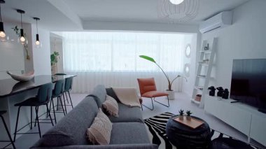 Şık bir açık-konsept oturma odasında dekoratif yastıklı gri bir kanepe, bar tabureli parlak siyah bir mutfak tezgahı, kahverengi deri bir sandalye ve beyaz bir eğlence merkezi bulunur..