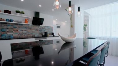 Parlak siyah bir adanın olduğu çağdaş bir mutfak, mavi kartonpiyer kaplama, beyaz dolap, dekorlu açık raflar, kolye aydınlatma, ve parlak siyah bir kaput.