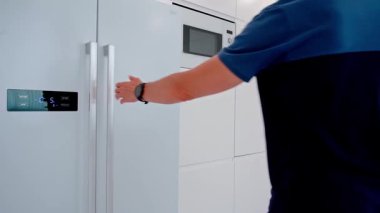 Donanma mavisi polo ve kol saati takan bir adam modern mutfakta dijital ısı göstergesi olan beyaz bir buzdolabını yan yana açar.