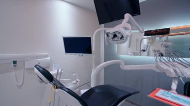 Siyah yastıklı modern dişçi koltuğu, ayarlanabilir kolluklar, monte monitörler, tepe lambası, tıbbi gereçler, ve parlak ışıklandırmalı temiz organize diş kliniğinde ergonomik tasarım