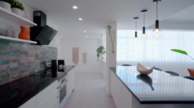 Parlak siyah tezgahı olan modern bir mutfak, mavi seramikli bir seramik, dekorlu açık raflar, askılı kolye ışıkları, ve parlak bir iç mekanda makrome duvar resimleri.