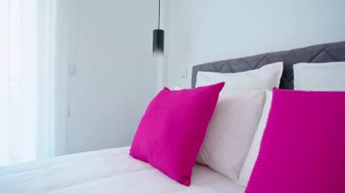 Modern yatak odasında düzgün yapılmış, pembe yastıklı, koyu renk yastıklı, katlanmış havlular, katlanmış perdeler, kolye ışıkları ve yumuşak doğal ışık altında yapılmış gardıroplar var..