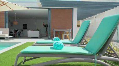 Temiz turkuaz havlular, parlak bir havuzun yanındaki güneşli yataklara davetkar bir şekilde uzanır. Arka planda modern, açık bir villa bulunur. Rahatlama ve tatil havası vaat eder.