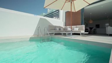 Turkuaz su, modern bir villaya bitişik özel bir yüzme havuzunda parıldıyor, beyaz bir kanepe, yastıklar ve güneş şemsiyesi ile tamamlanmış, rahatlama ve rahatlama için huzurlu bir ortam yaratıyor.
