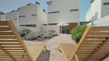 Modern beyaz evler arka bahçeyi ahşap güneşlikler, çakıllar, küçük ağaçlar ve çalılarla çevreler. Rahatlatıcı ve şık bir açık hava alanı yaratırlar.