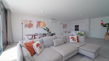 Turuncu ve bej yastıklı gri kanepe geniş oturma odasında dinleniyor, doğal ışığa sahip ve yemek alanı ile kusursuz bir şekilde birbirine bağlı.