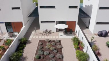 Modern beyaz konut binasının birden fazla görüntüsü, bitkilerle, açık mobilyalarla, bisikletlerle ve çatıda bir terası olan rahat bir bahçeyi gösteriyor.